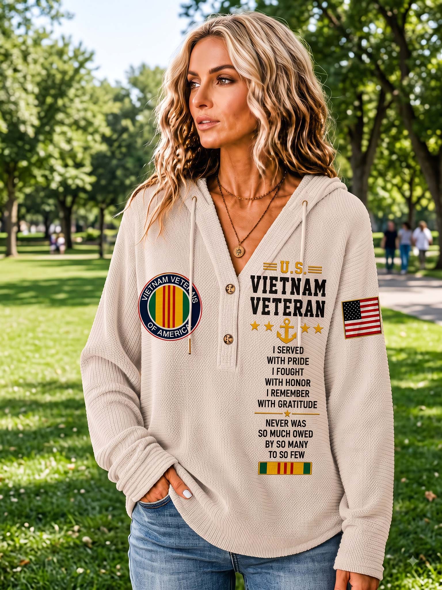 A woman wearing a beige ribbed hoodie with a 'Vietnam Veteran' graphic print on the front, standing in a sunny park with trees in the background.