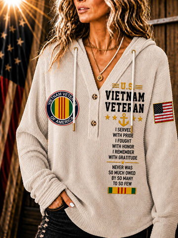 A woman wearing a beige ribbed hoodie with 'Vietnam Veteran' text, military-style patches, and an American flag graphic on the sleeve, posed against a rustic wooden background with a blurred American flag.