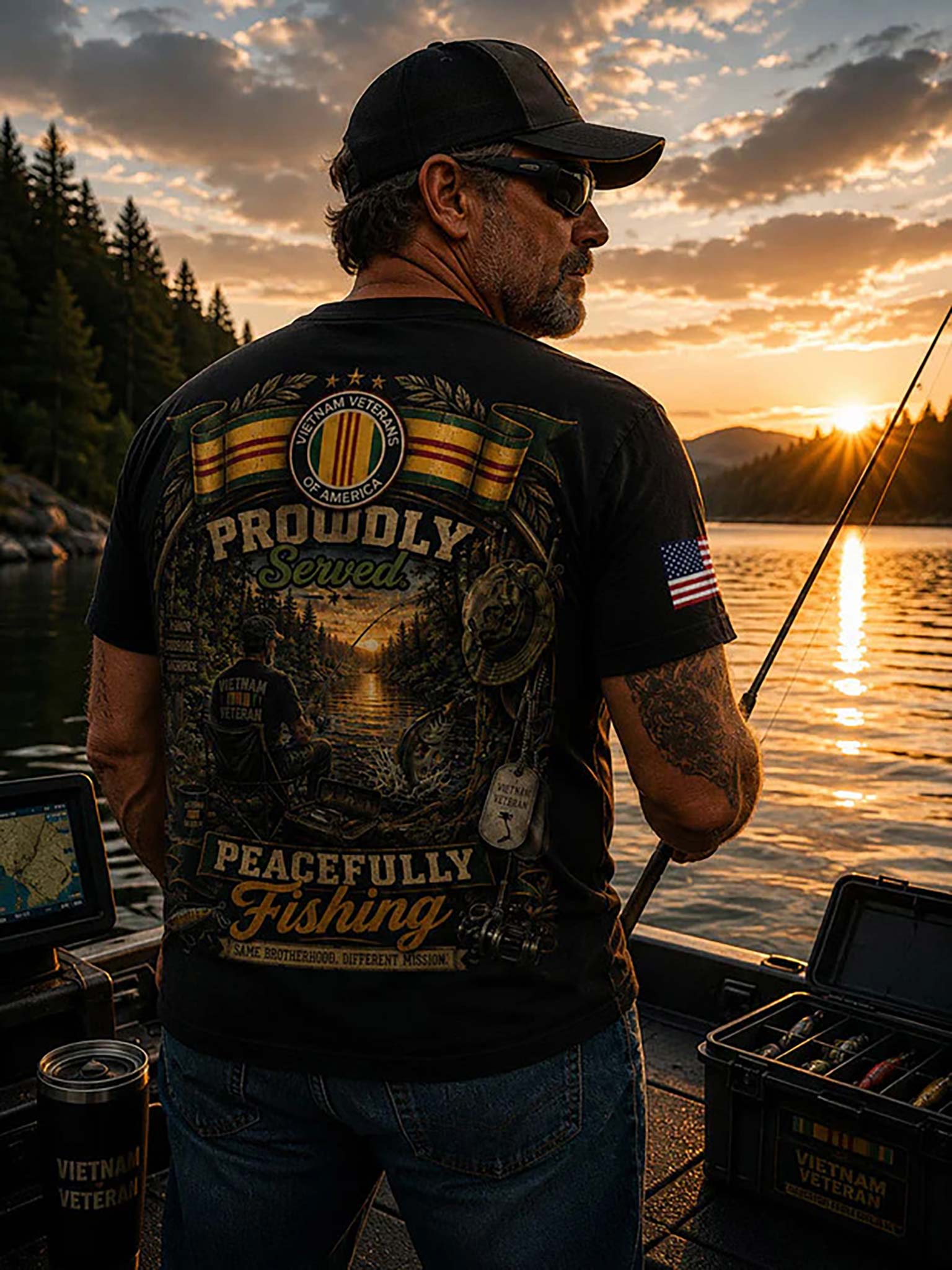 A man wearing a black baseball cap and sunglasses stands on a boat at sunset, facing away from the camera. He is wearing a black graphic t-shirt with a large back print that reads 'Proudly Served Peacefully Fishing' and an American flag patch on the sleeve.