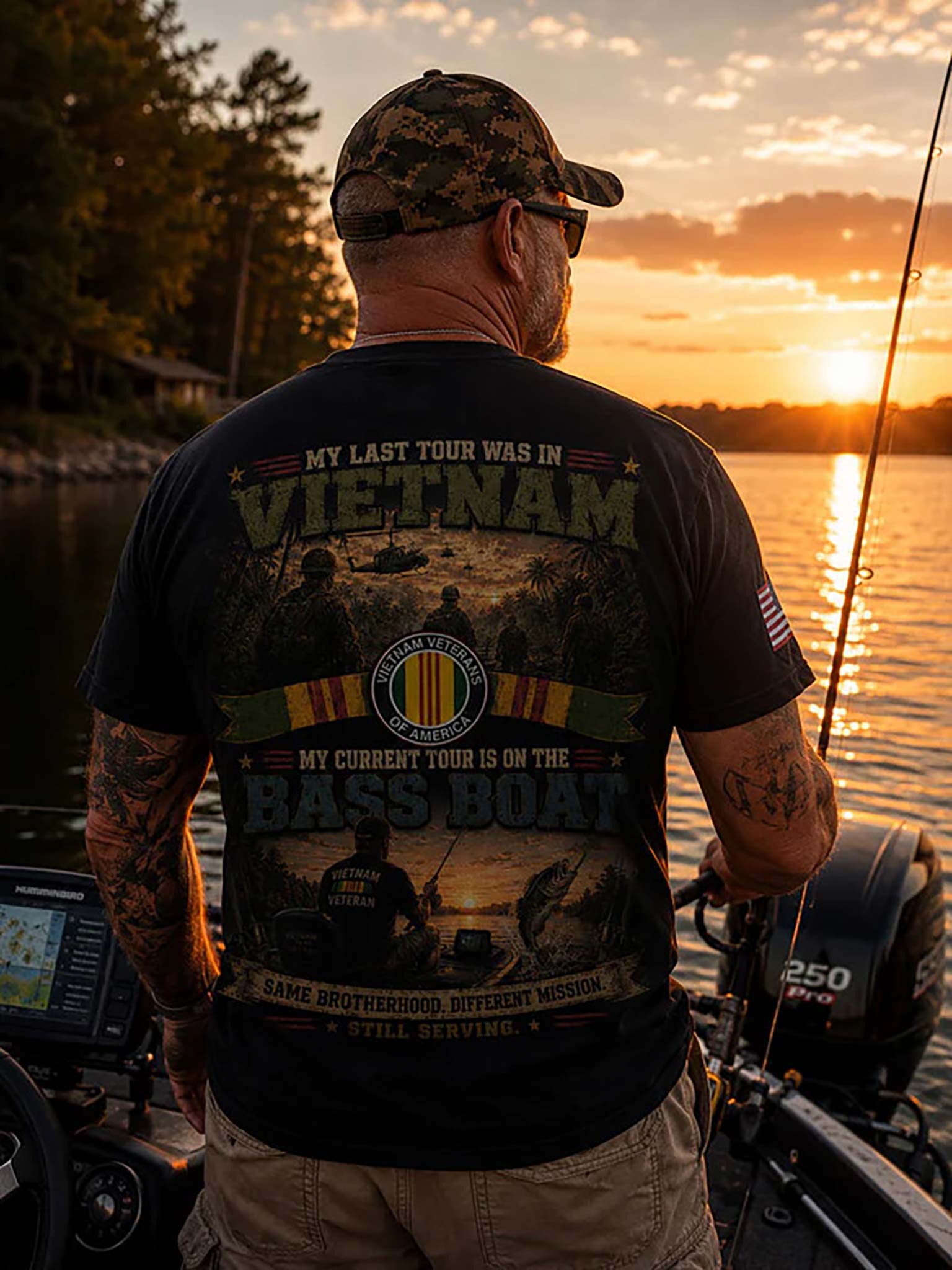 A man seen from behind wearing a black graphic t-shirt and a camouflage cap on a boat during sunset. The back of the t-shirt features a large military-themed graphic with the text 'MY LAST TOUR WAS IN VIETNAM' and 'MY CURRENT TOUR IS ON THE BASS BOAT'.