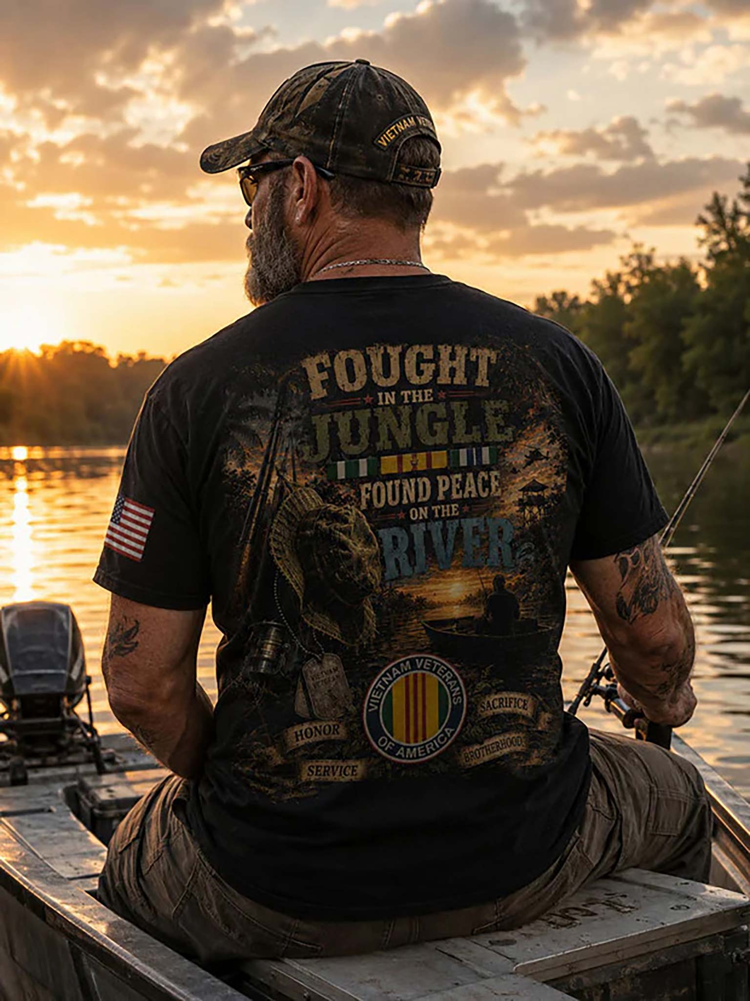 A man sitting in a boat on a lake at sunset, viewed from behind, wearing a black graphic t-shirt with a military-themed design that reads 'FOUGHT IN THE JUNGLE TO FIND PEACE ON THE RIVER'.