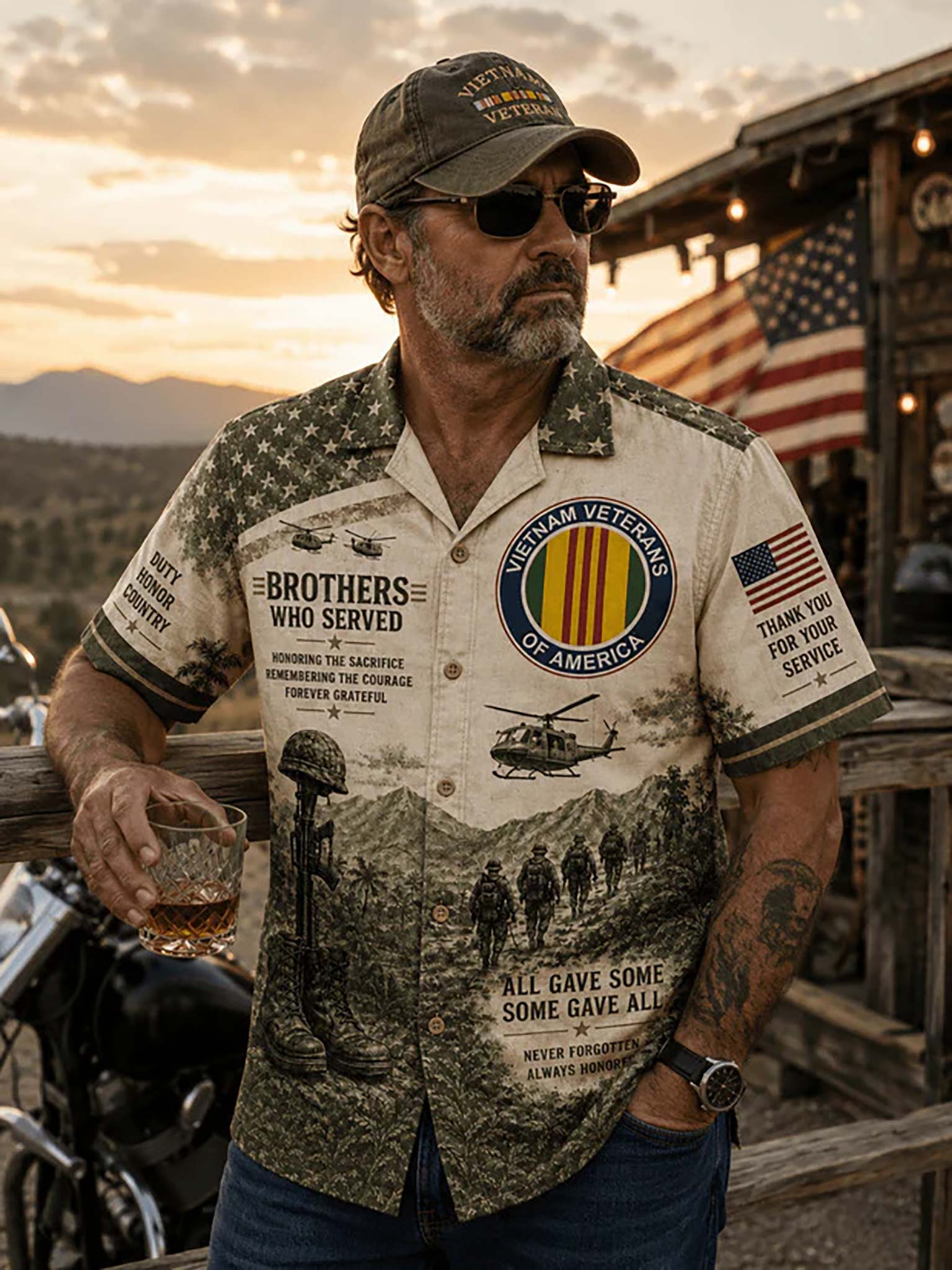 A man wearing a military-themed short-sleeve button-up shirt with 'Veterans Who Served' graphics, standing outdoors at sunset near a rustic wooden structure and motorcycle.