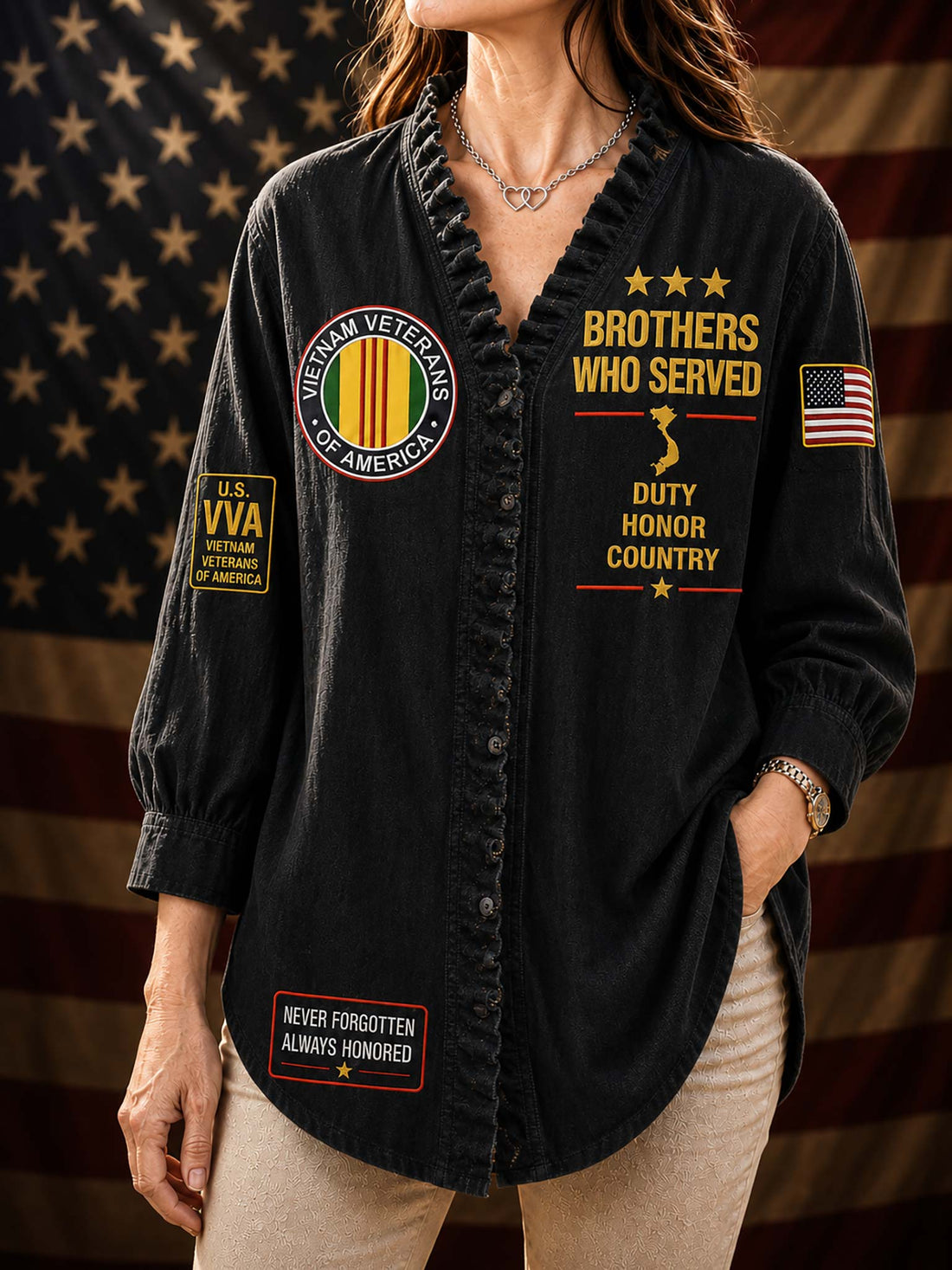A woman wearing a black long-sleeve button-front shirt with patriotic military-themed graphics, including 'Brothers Who Served,' 'Veterans of America,' and an American flag patch, posed against a blurred American flag background.
