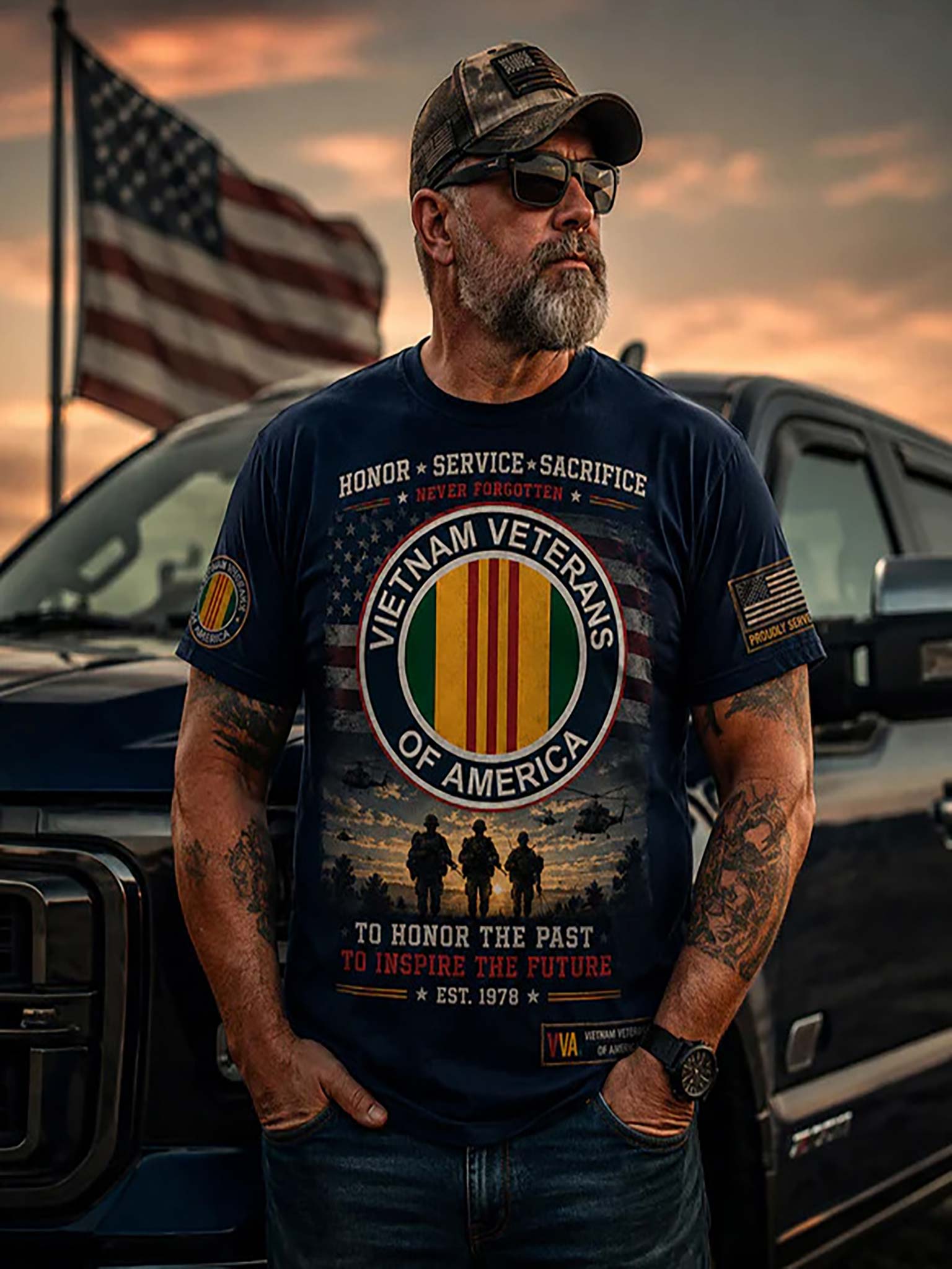 A man wearing a navy blue graphic t-shirt with a Vietnam Veterans of America design, standing in front of a black pickup truck and an American flag at sunset.