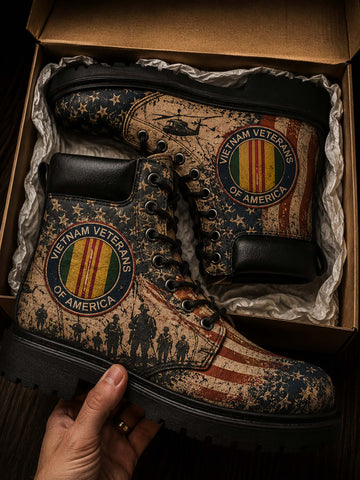 A pair of distressed military-style boots with a patriotic American flag and stars-and-stripes print, displayed inside a cardboard box with a hand holding one boot. The boots feature circular 'Vietnam Veterans of America' emblems and a helicopter graphic.