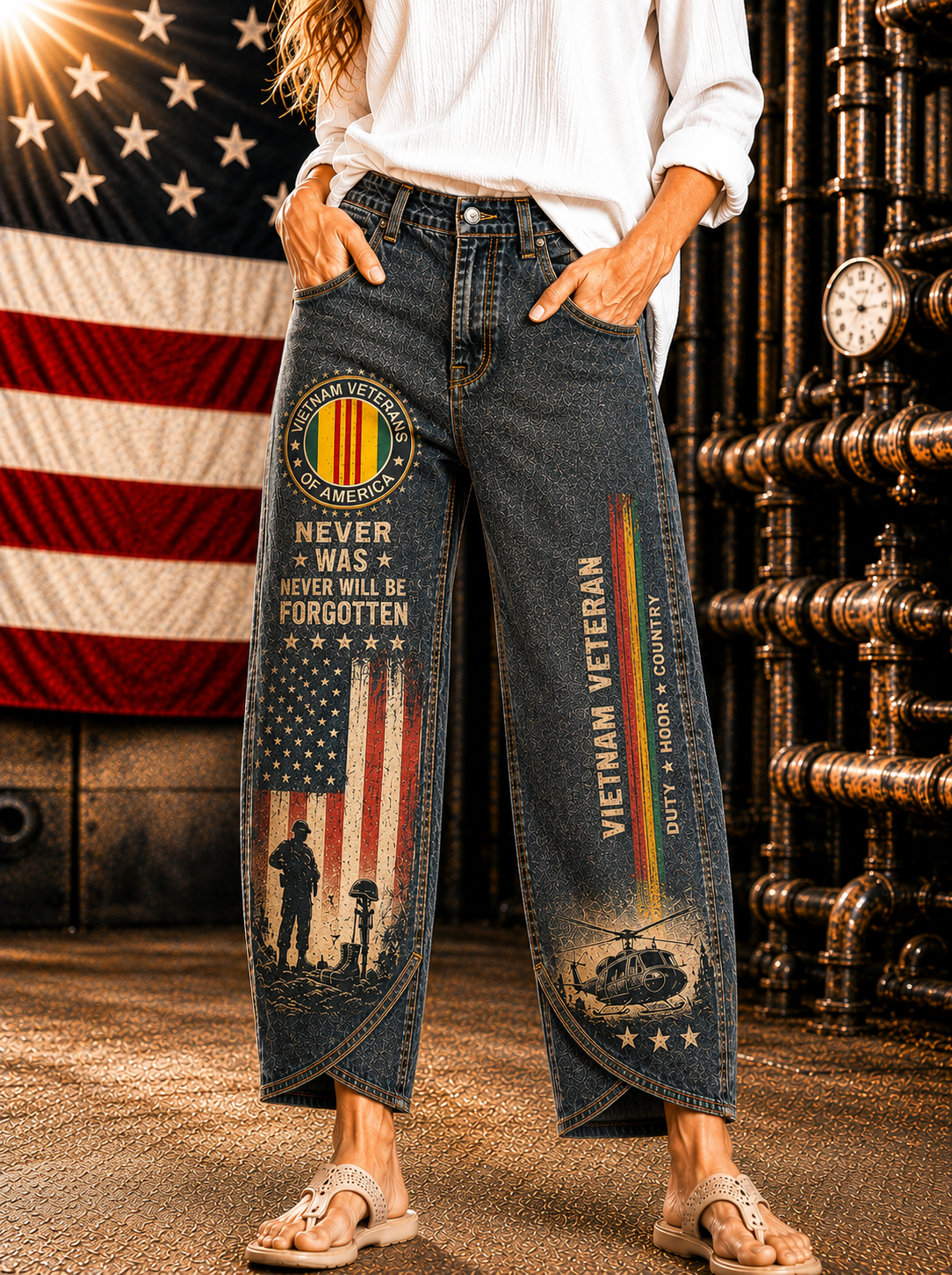 A person wearing dark blue cropped jeans with patriotic military-themed prints, including an American flag, 'Never Was Never Will Be Forgotten' emblem, and 'Vietnam Veteran' text, standing in front of an American flag and industrial pipe structure.