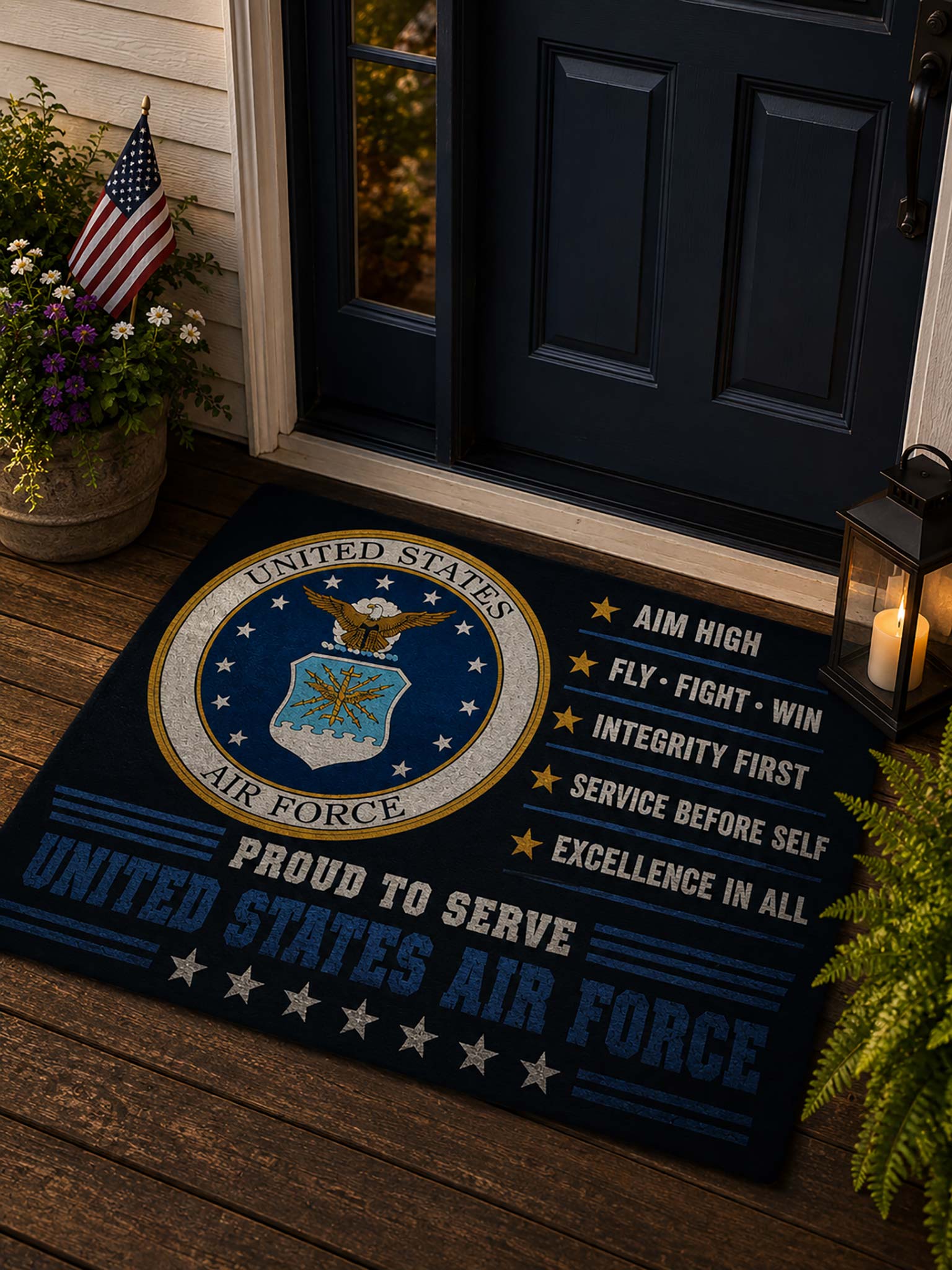 A dark navy blue doormat featuring the United States Air Force seal and the words 'United States Air Force' and 'Proud to Serve,' placed in front of a dark blue front door on a wooden porch with potted plants and a lantern nearby.