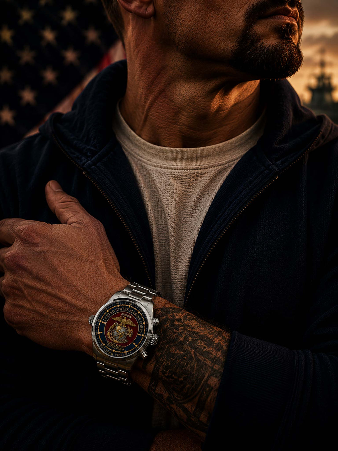 A stainless steel military-style wristwatch with a detailed United States Marine Corps themed dial, featuring the USMC emblem, 'United States Marine Corps' text, and a red, gold, and blue face. The watch is set against a blurred American flag background on a wooden surface.