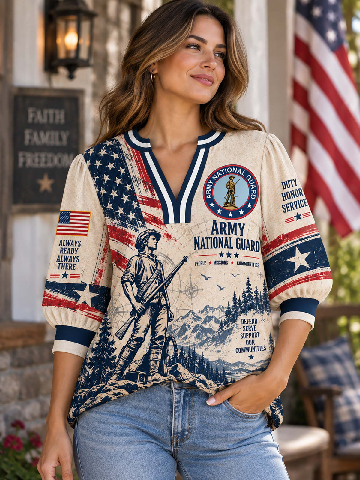 A woman wearing a patriotic Army National Guard graphic t-shirt with blue jeans, standing on a porch with an American flag in the background.