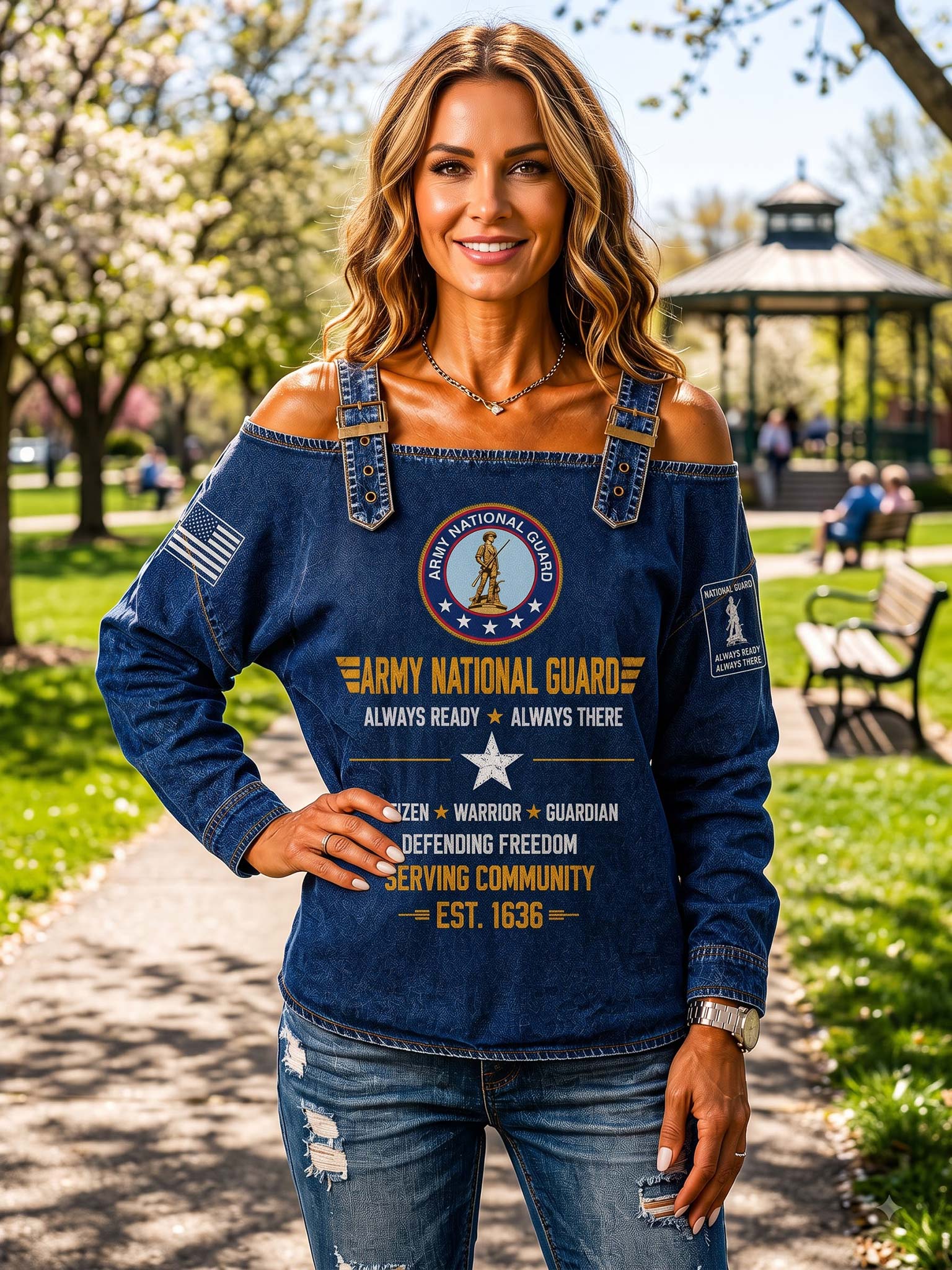 A woman stands outdoors in a park wearing a blue off-the-shoulder long-sleeve top with Army National Guard graphics and distressed blue jeans.