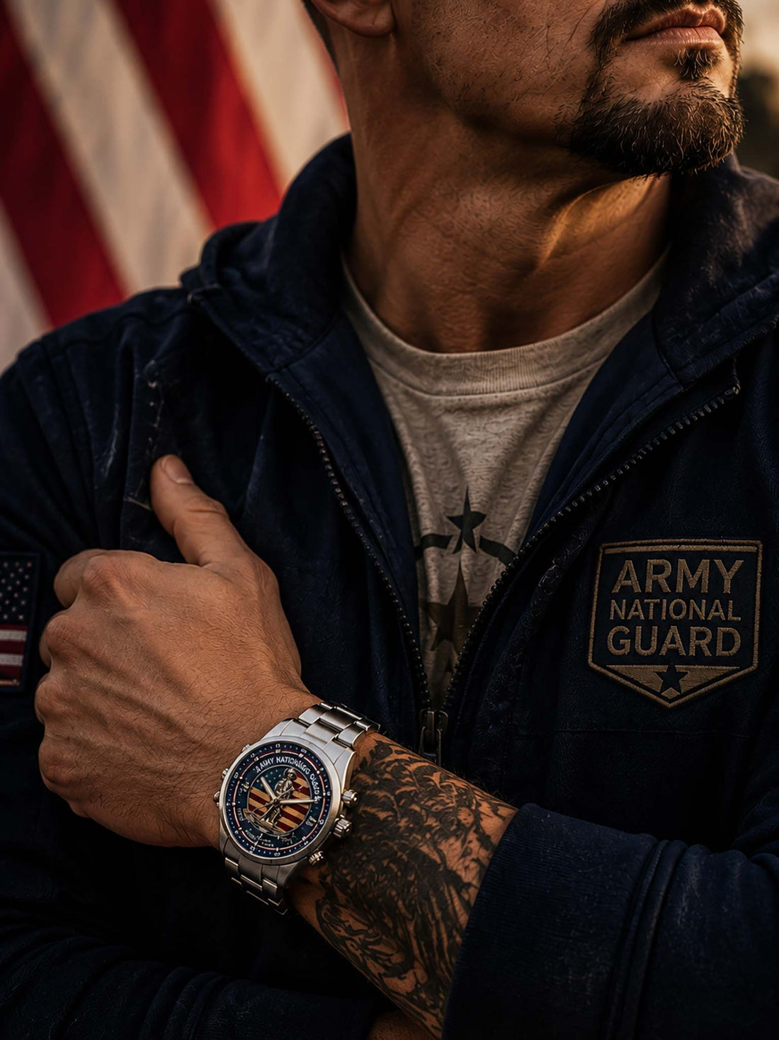 A stainless steel military-themed watch with a round silver-tone case and link bracelet, featuring a detailed dial with the words 'ARMY NATIONAL GUARD,' an American flag background, a central soldier statue, and text including 'DEFEND • SERVE • PROTECT' and 'UNITED STATES.' The watch is photographed on a wooden surface with a blurred American flag backdrop.