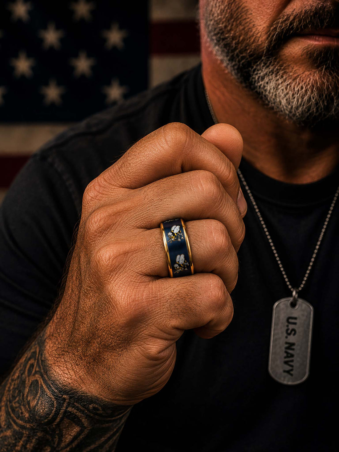 Two navy blue Seabees metal rings with gold trim and bee motifs, one engraved with 'SEABEES' on the inside, displayed against a blurred American flag background.