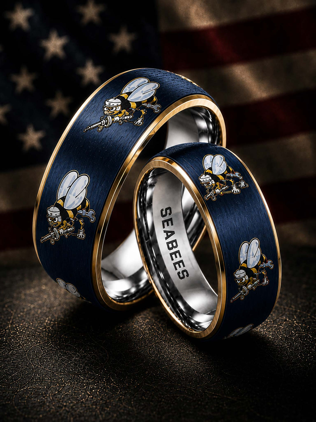 Two navy blue Seabees metal rings with gold trim and bee motifs, one engraved with 'SEABEES' on the inside, displayed against a blurred American flag background.