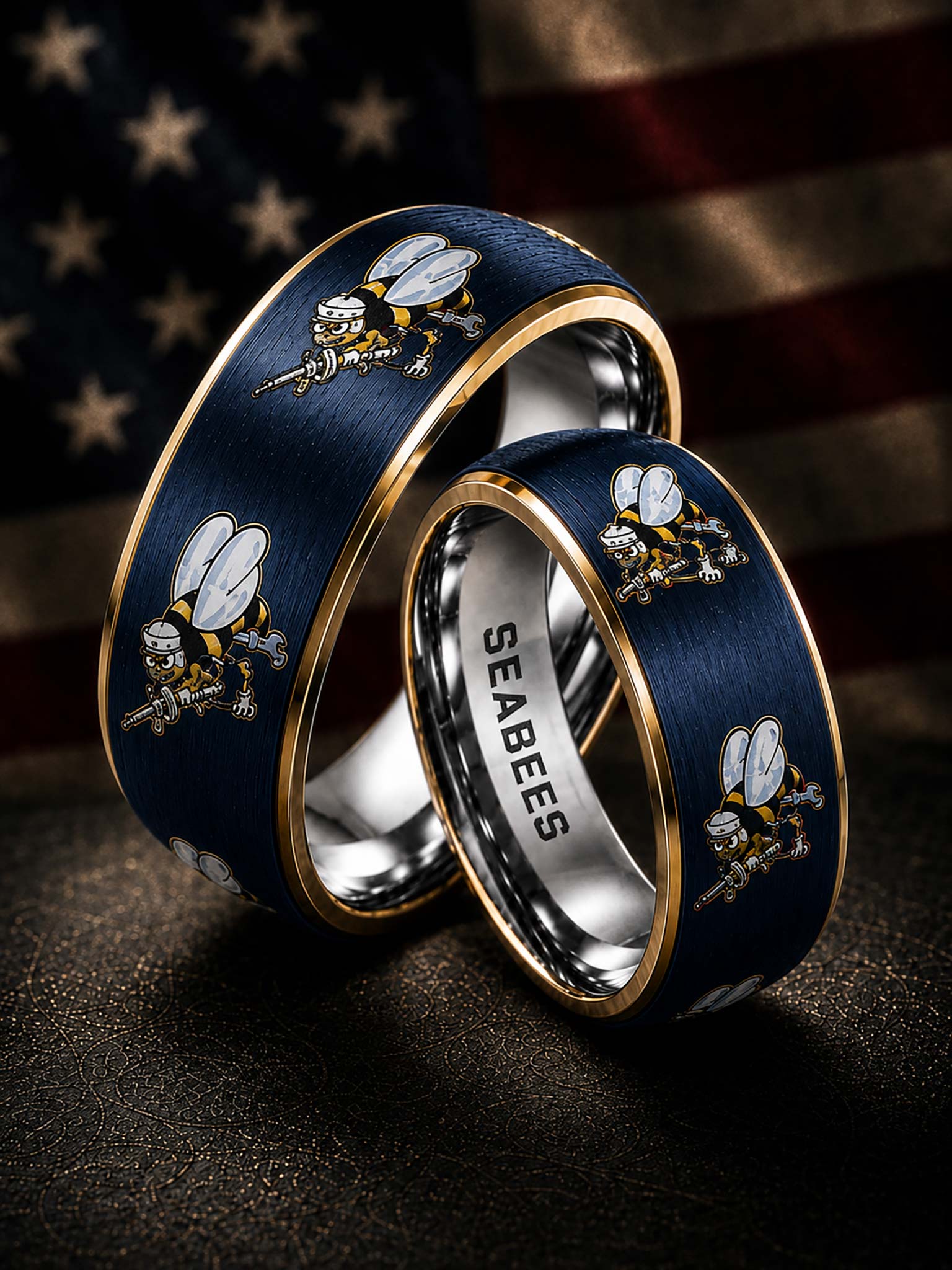 Two navy blue Seabees metal rings with gold trim and bee motifs, one engraved with 'SEABEES' on the inside, displayed against a blurred American flag background.