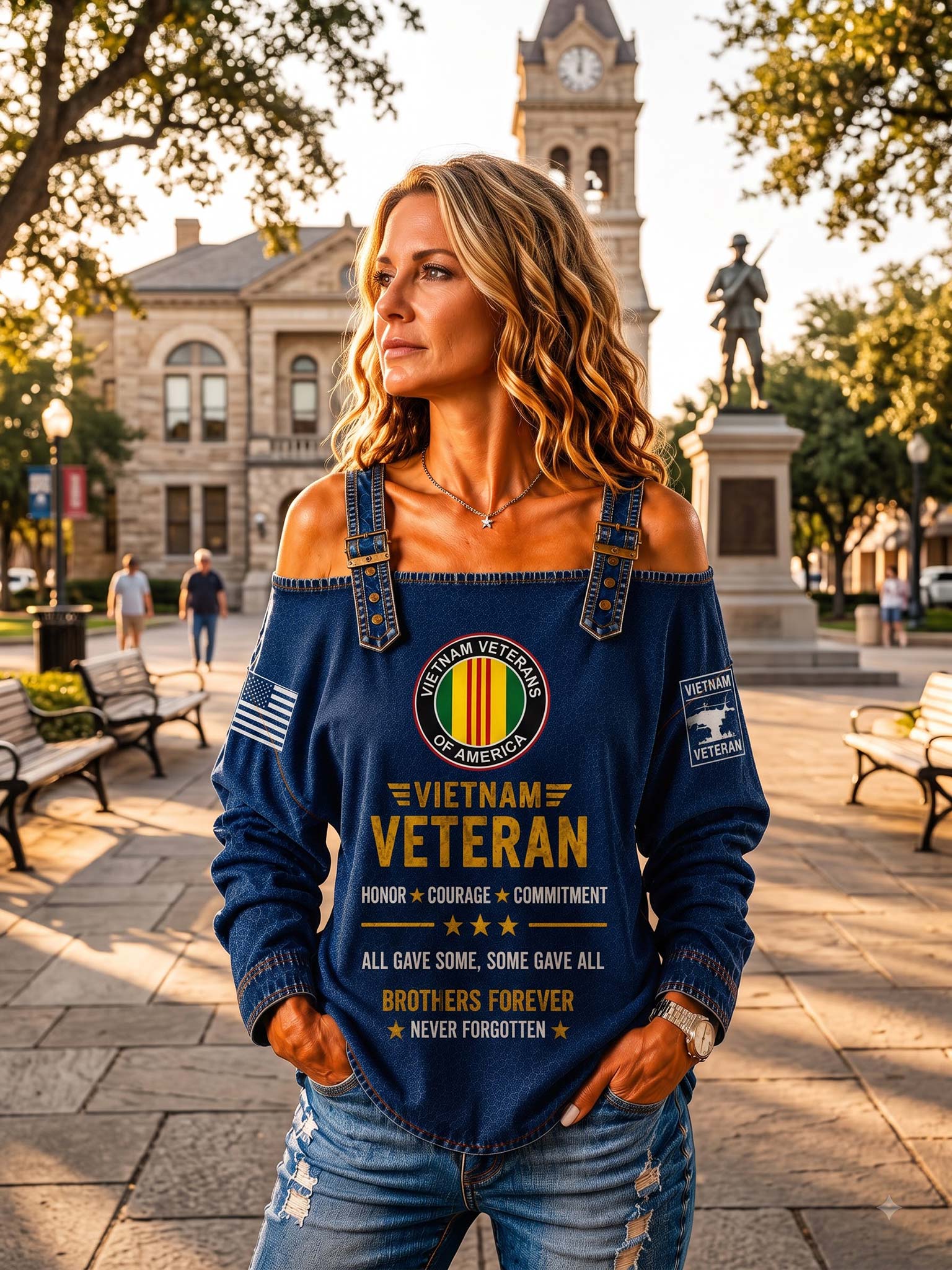 A woman wearing a navy blue off-the-shoulder long-sleeve sweatshirt with a 'Vietnam Veteran' graphic print, standing in a sunny outdoor park plaza with a historic stone building in the background.